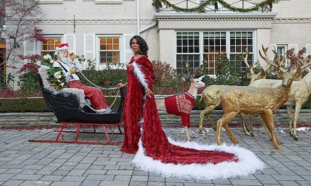 Ann Kaplan Mulholland in a red dress and cape next to a Christmas installation of Santa in his sled with reindeer.
