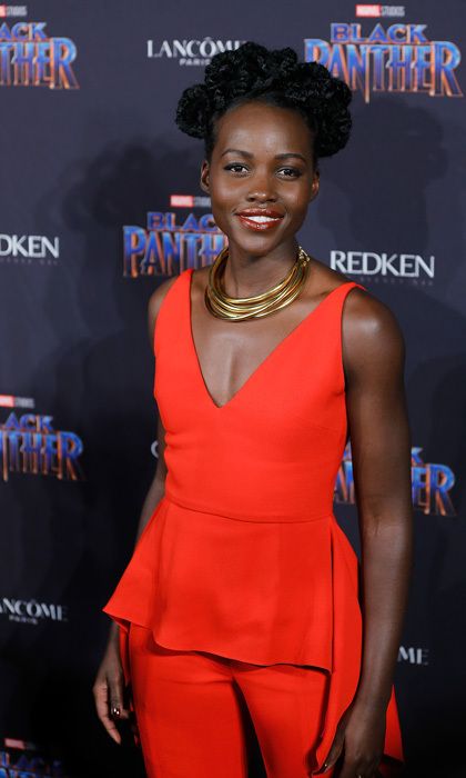 Lupita Nyong'o hair in a pretzel do seen in a red outfit and stacked gold rings neclace.